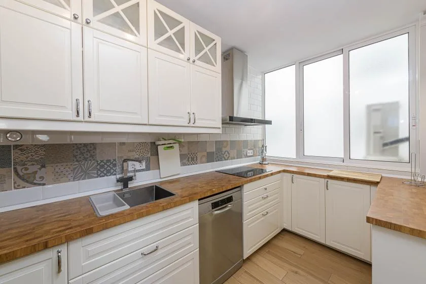 Una cocina luminosa con gabinetes blancos, encimeras de madera y ventanas grandes.