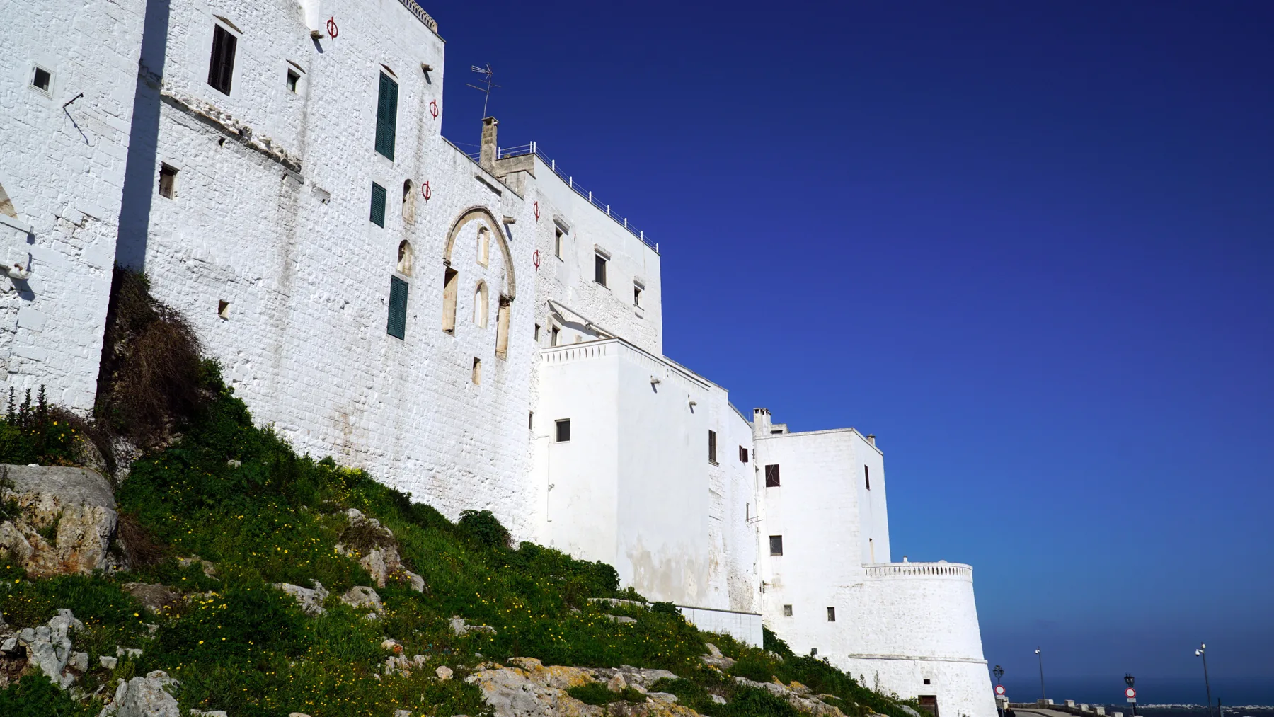 Edificios blancos se alzan sobre una ladera rocosa y verde bajo un cielo azul despejado.
