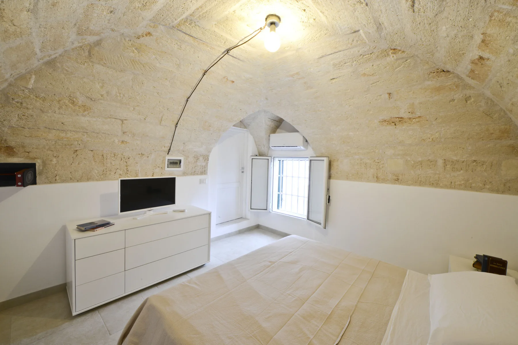 Un dormitorio con una cama, una cómoda y una ventana abierta bajo un techo abovedado.