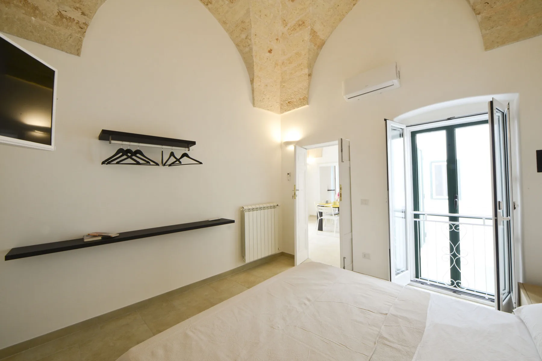 Un dormitorio luminoso con cama, televisor en la pared y puertas abiertas al balcón.