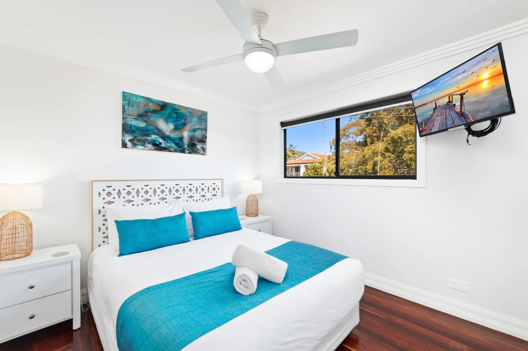 Un dormitorio luminoso con ropa de cama blanca, detalles turquesa y una ventana con vista a los árboles.