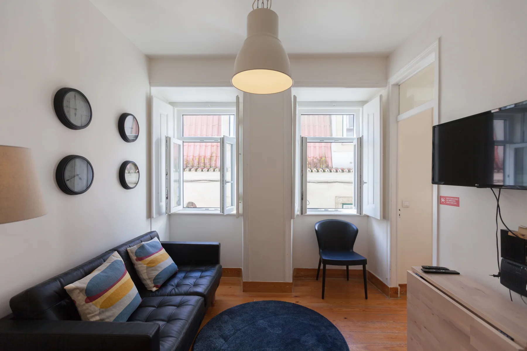 Sala de estar con un sofá oscuro, una silla y dos ventanas luminosas.