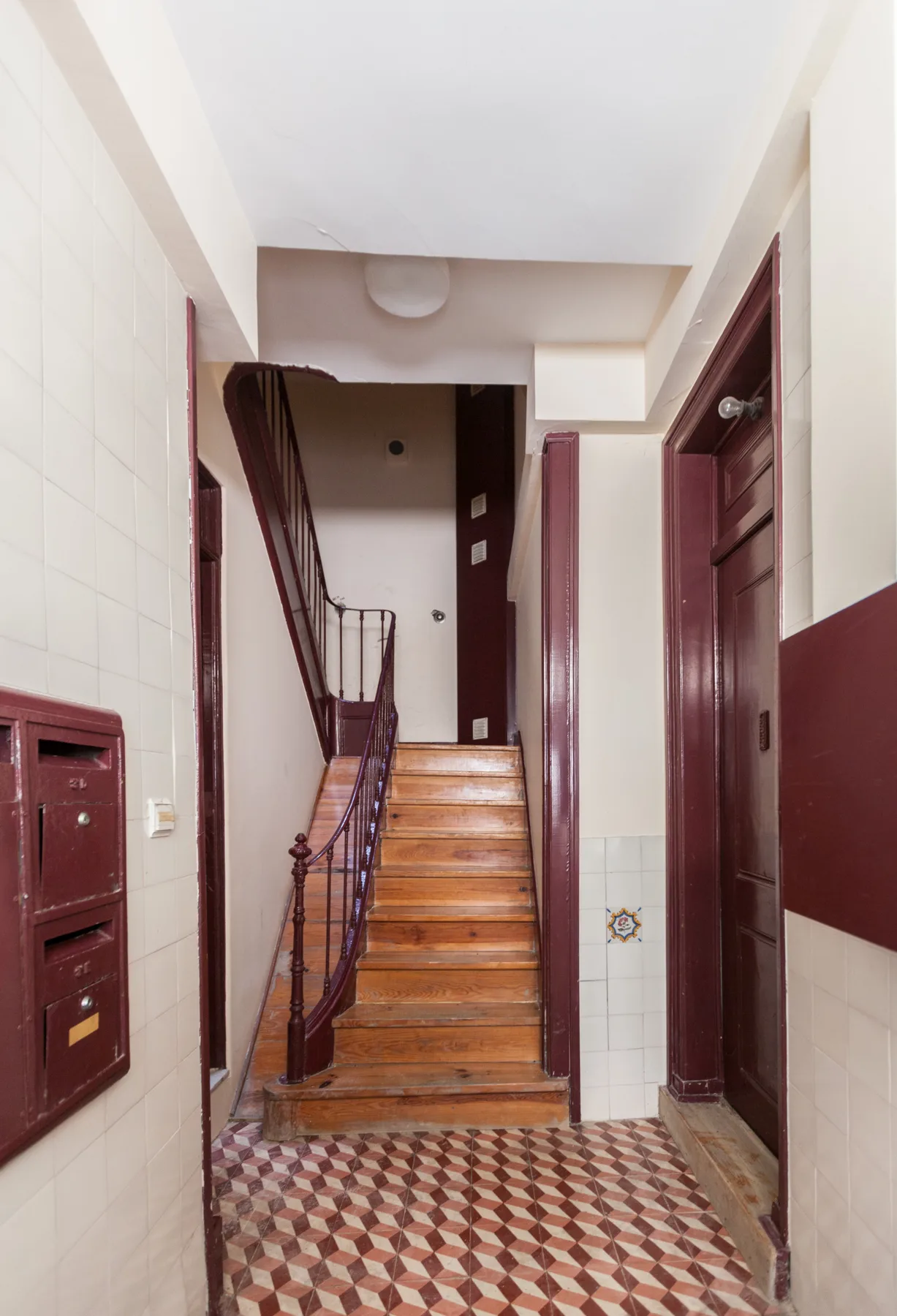 Un pasillo interior de baldosas conduce a una escalera de madera con barandilla oscura.