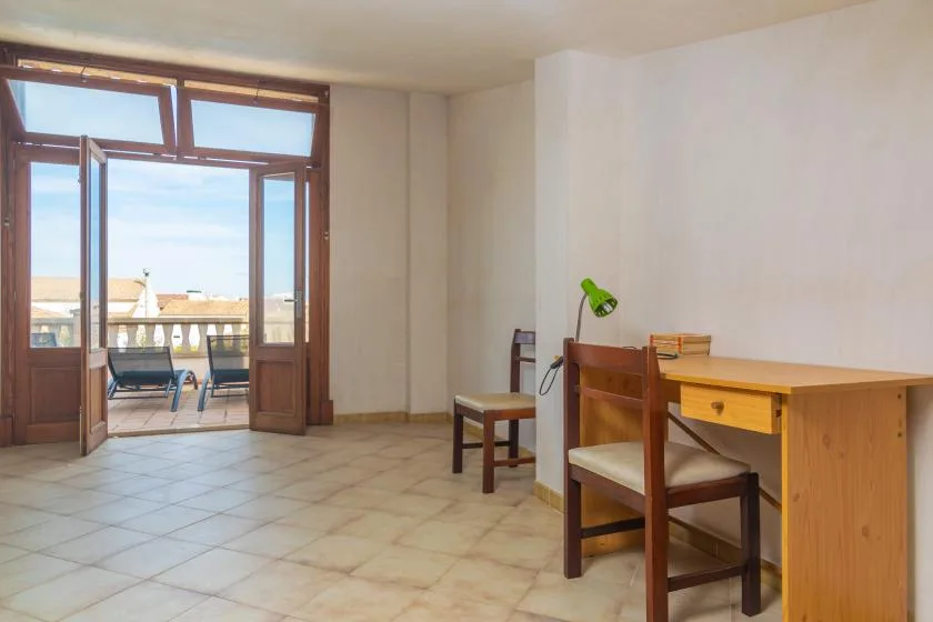 Una habitación con suelo de baldosas, escritorio de madera y asientos junto a puertas abiertas al balcón.