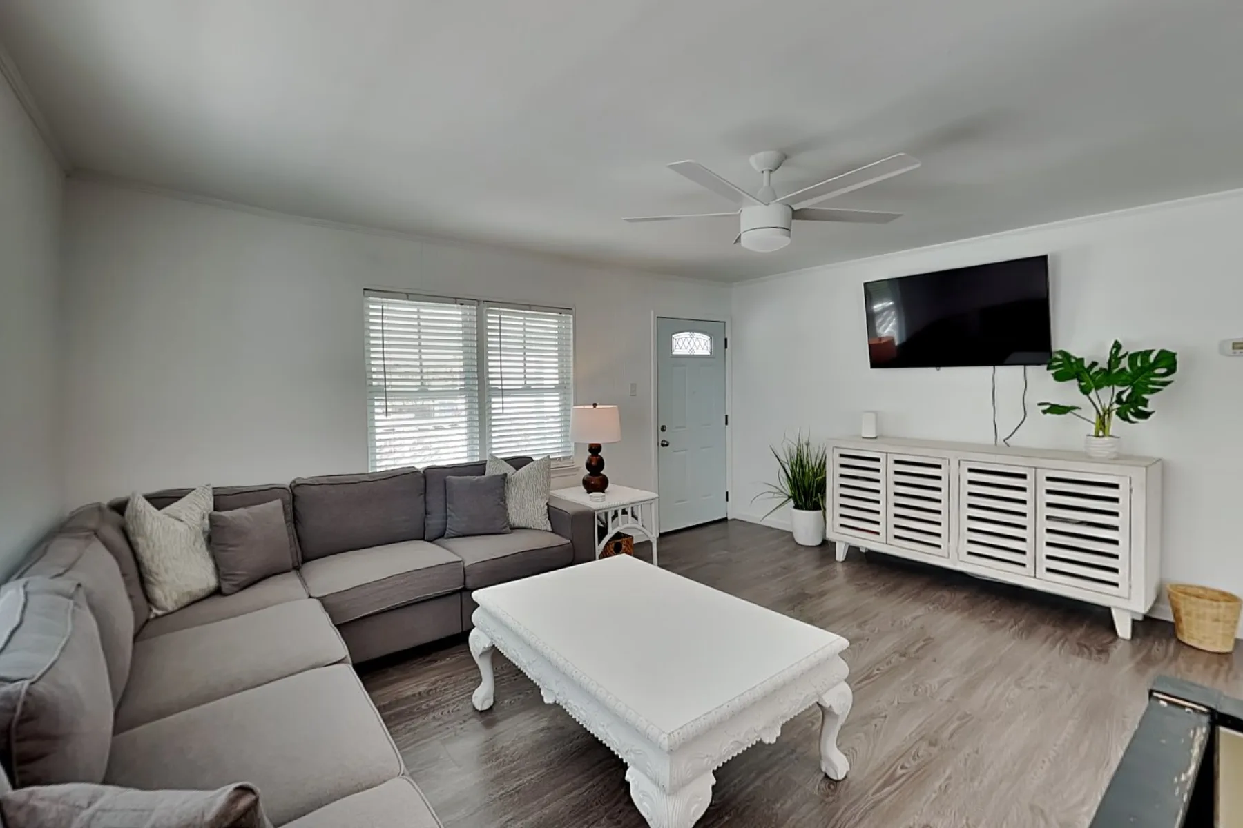 A bright living room with sectional seating, a white coffee table, and a wall-mounted television.