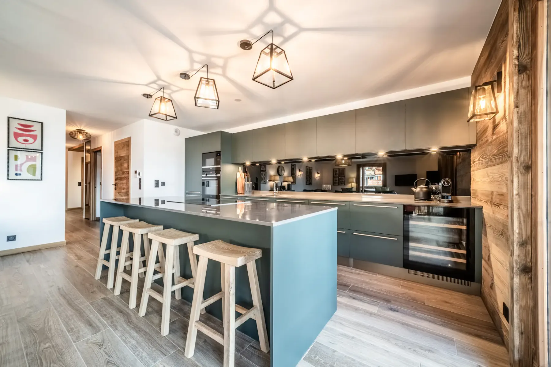 Modern kitchen with gray cabinets, a central island with bar stools, wooden dining table, pendant lighting, and large windows overlooking a balcony and scenic view.
