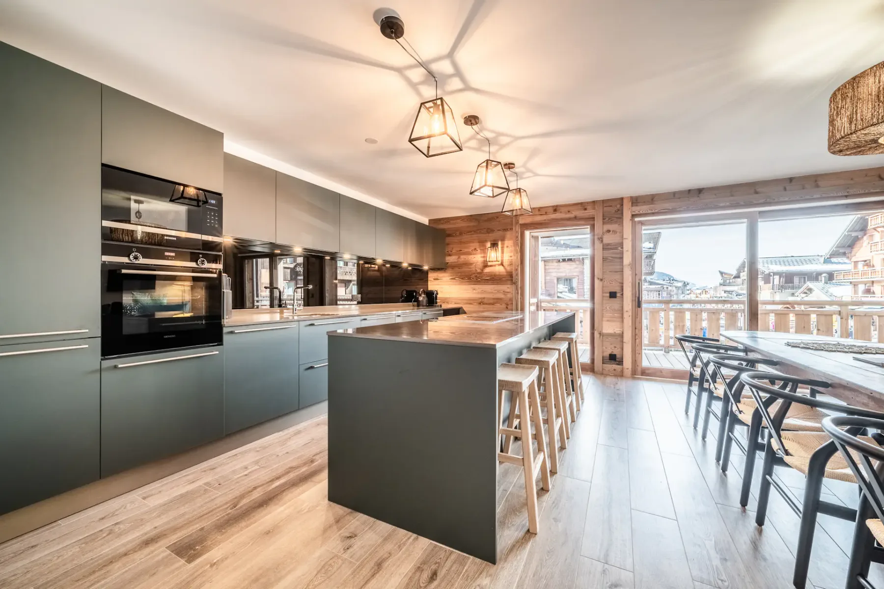 Modern kitchen with gray cabinets, a central island with bar stools, wooden dining table, pendant lighting, and large windows overlooking a balcony and scenic view.