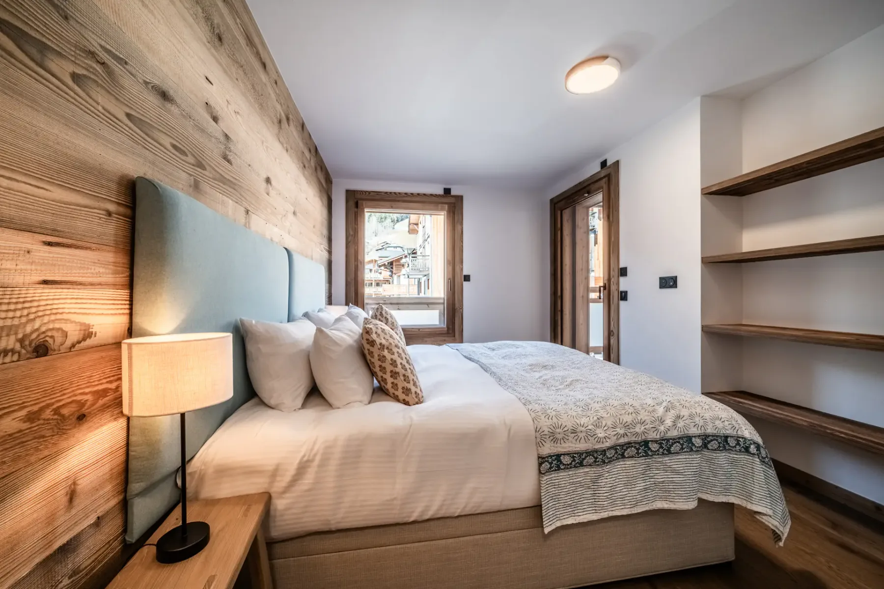 Cozy bedroom with a double bed, wooden walls, a bedside lamp, and built-in shelves. A window and glass door are visible, letting in natural light.