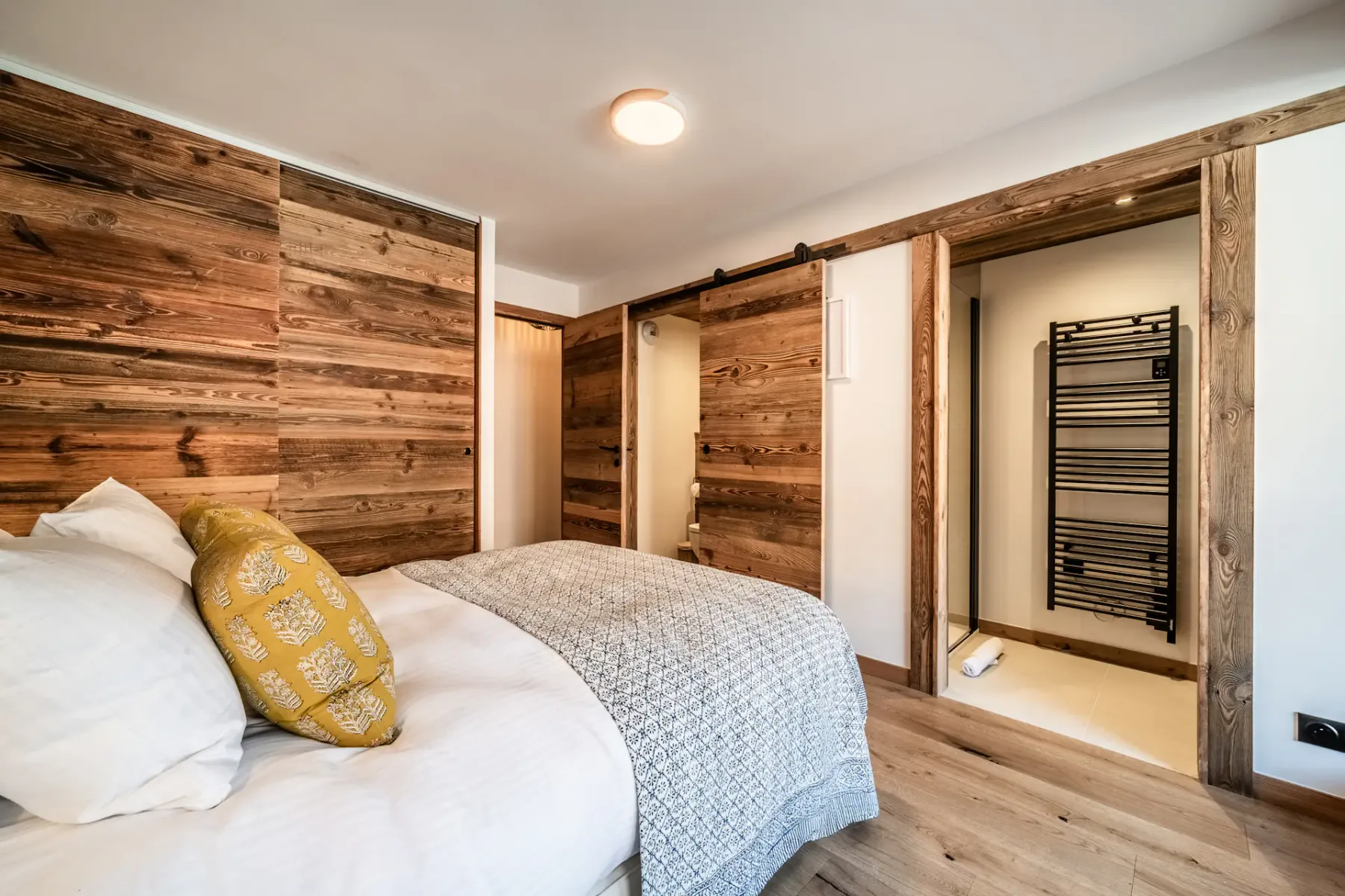 Cozy bedroom with a double bed, wooden walls, and large window doors opening to a balcony with a view of neighboring buildings.
