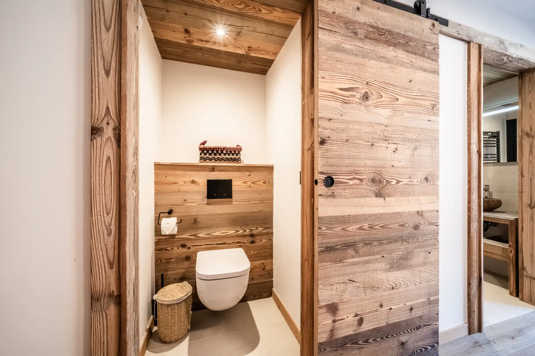 A modern bathroom with wooden sliding doors, a wall-mounted toilet, a toilet paper holder, and a wicker basket.