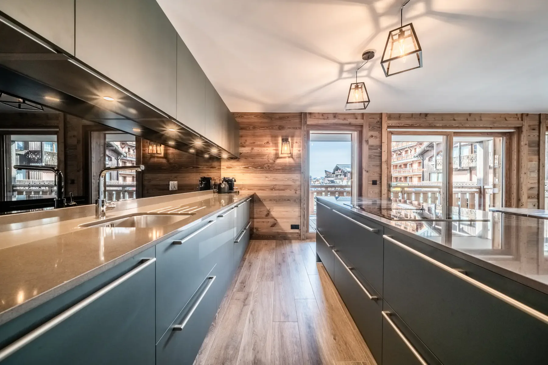 Modern kitchen with gray cabinets, a central island with bar stools, wooden dining table, pendant lighting, and large windows overlooking a balcony and scenic view.