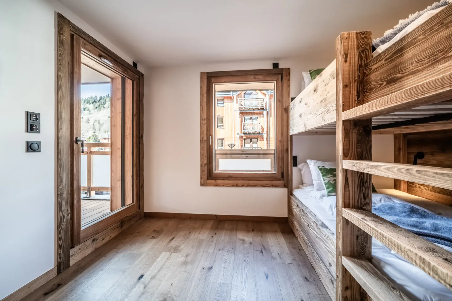 Wooden bunk beds in a bright room with a large window. The beds are neatly made with white linens. A ladder provides access to the upper bunks. The floor is wooden.