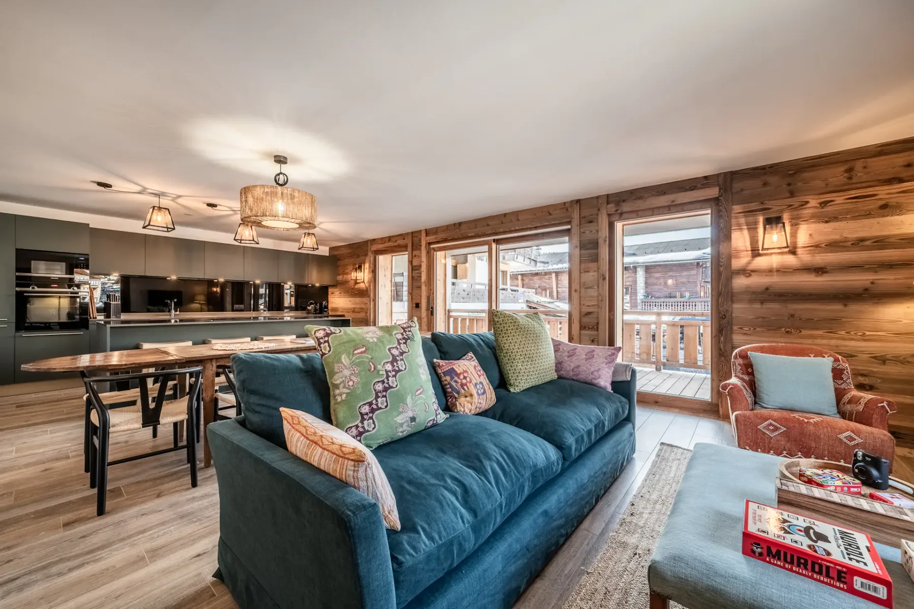 Living room with a teal sofa, orange armchair, and side tables. Wall art and lighting fixtures decorate white and wooden walls. A window provides natural light.