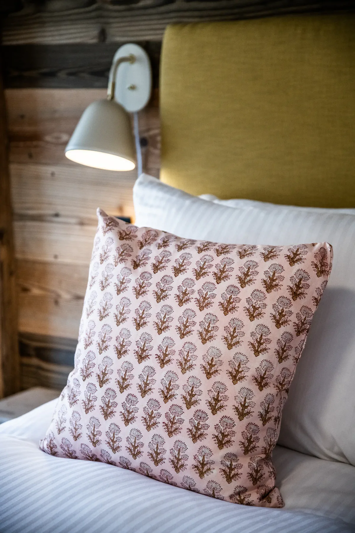 A patterned pillow rests on a white bed with a wooden wall and lit wall-mounted lamp in the background.
