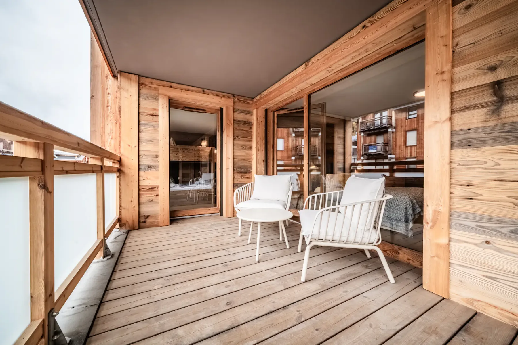 A wooden balcony with white chairs and small tables, overlooking neighboring buildings. Glass doors lead to the interior.