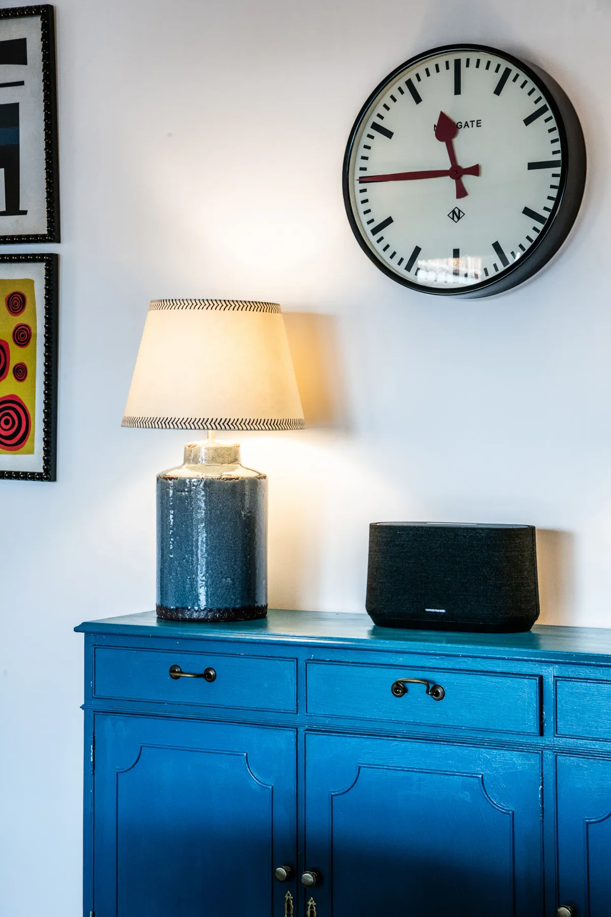 Close-up of a bed witBlue cabinet with a lamp and speaker on top, wall clock, and framed artworks hanging above.h a patterned pillow and a headboard. A lit lamp on a wooden wall is visible in the background.