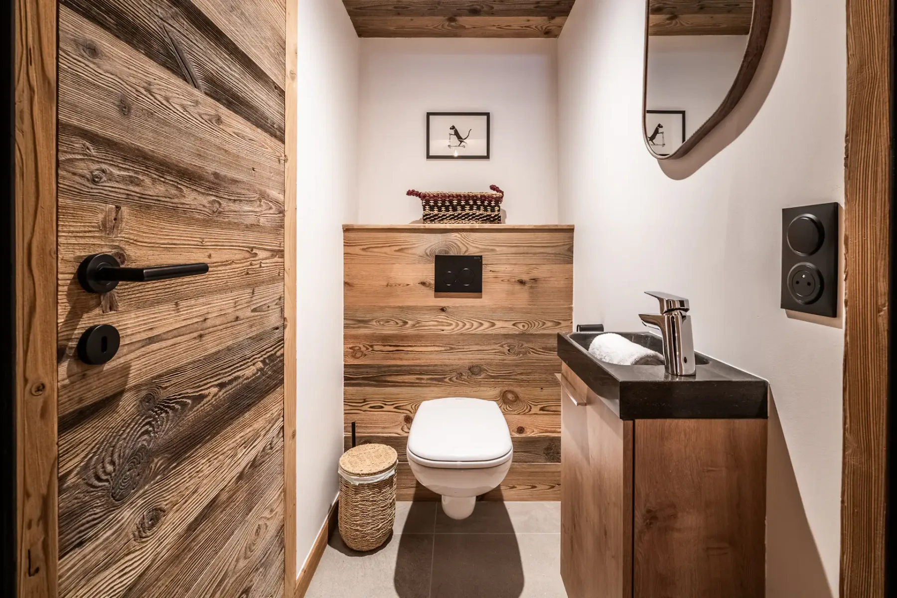 A small, modern bathroom with a wall-mounted toilet, wooden paneling, sink with a black countertop, circular mirror, and a decorative basket on the floor.
