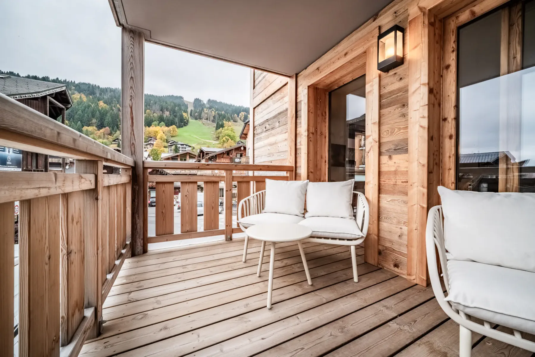 A wooden balcony with white chairs and small tables, overlooking neighboring buildings. Glass doors lead to the interior.
