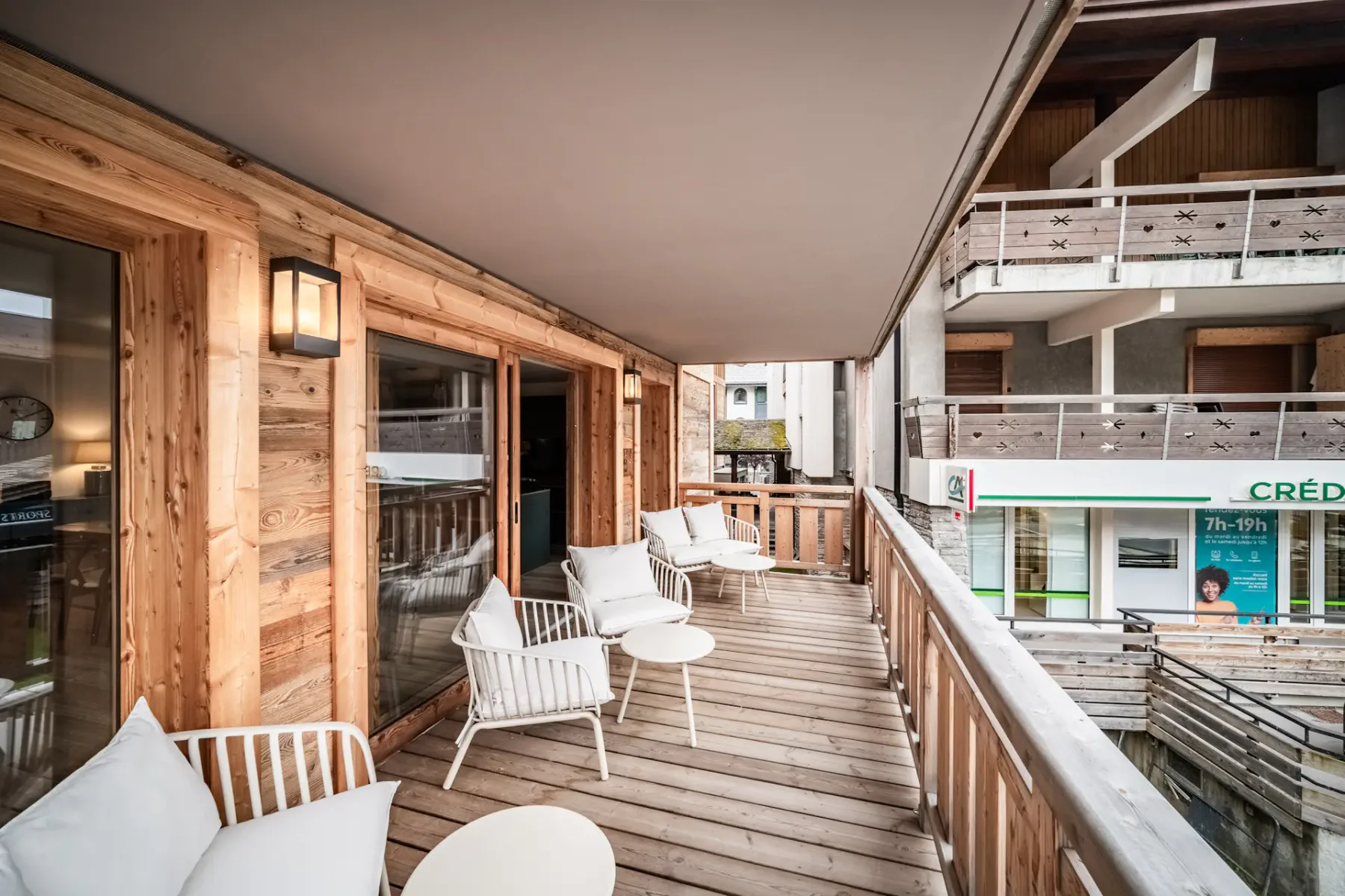 A wooden balcony with white chairs and small tables, overlooking neighboring buildings. Glass doors lead to the interior.