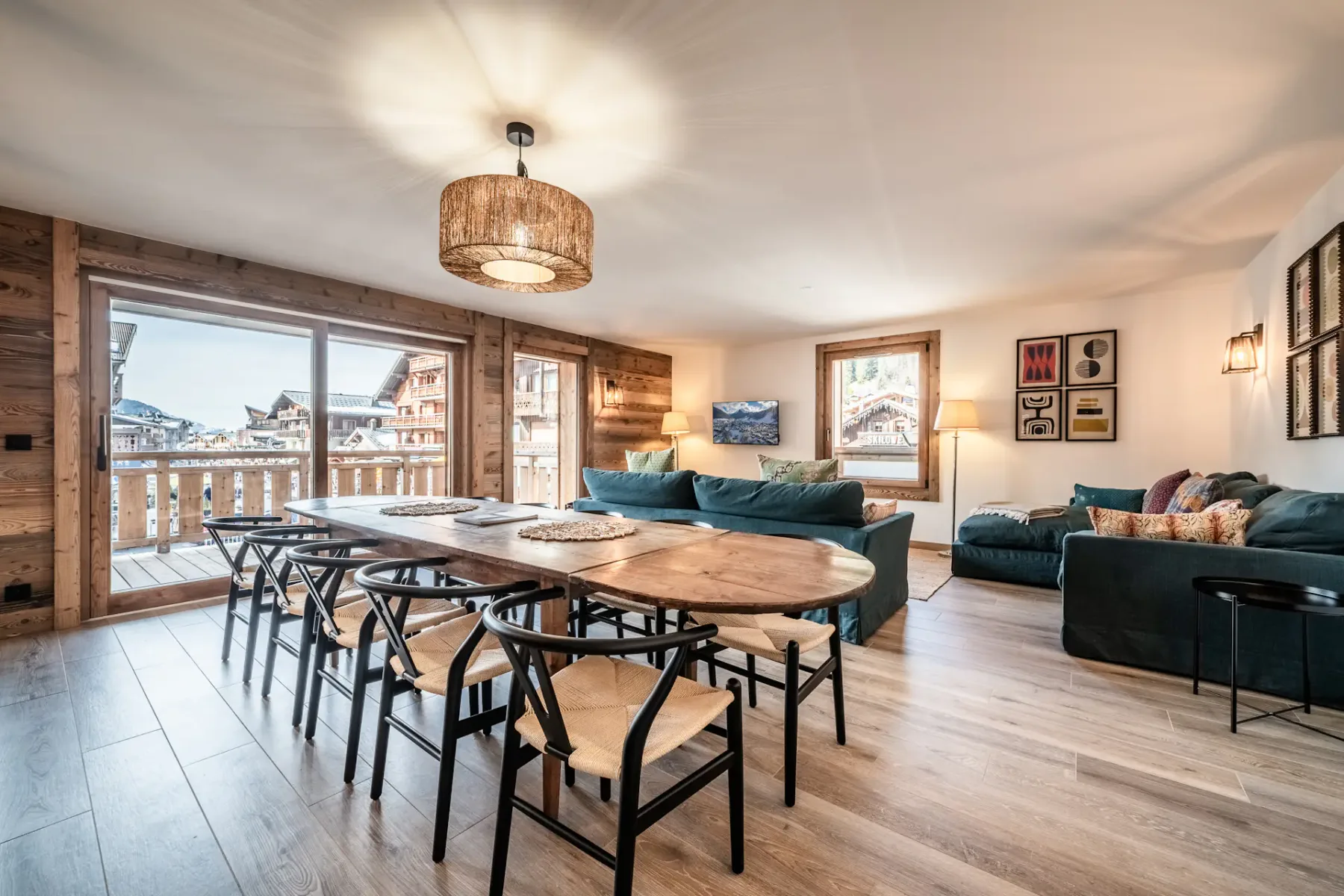 Modern kitchen with gray cabinets, a central island with bar stools, wooden dining table, pendant lighting, and large windows overlooking a balcony and scenic view.