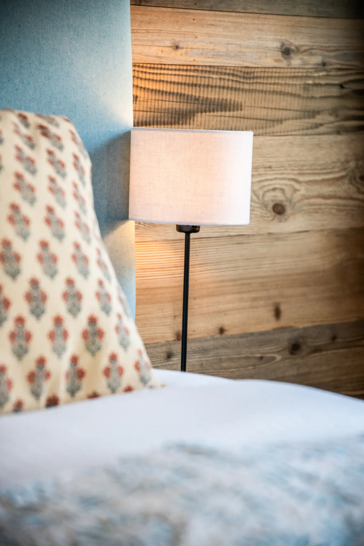 Close-up of a bed with a patterned pillow and a headboard. A lit lamp on a wooden wall is visible in the background.