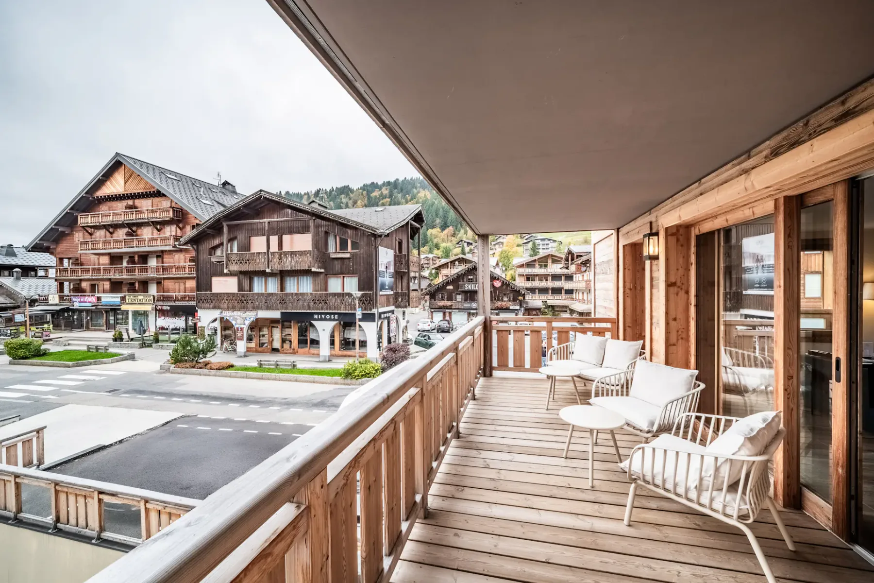 Wooden terrace overlooking a quaint mountain village with rustic buildings, a few people, and parked cars under a clear blue sky.