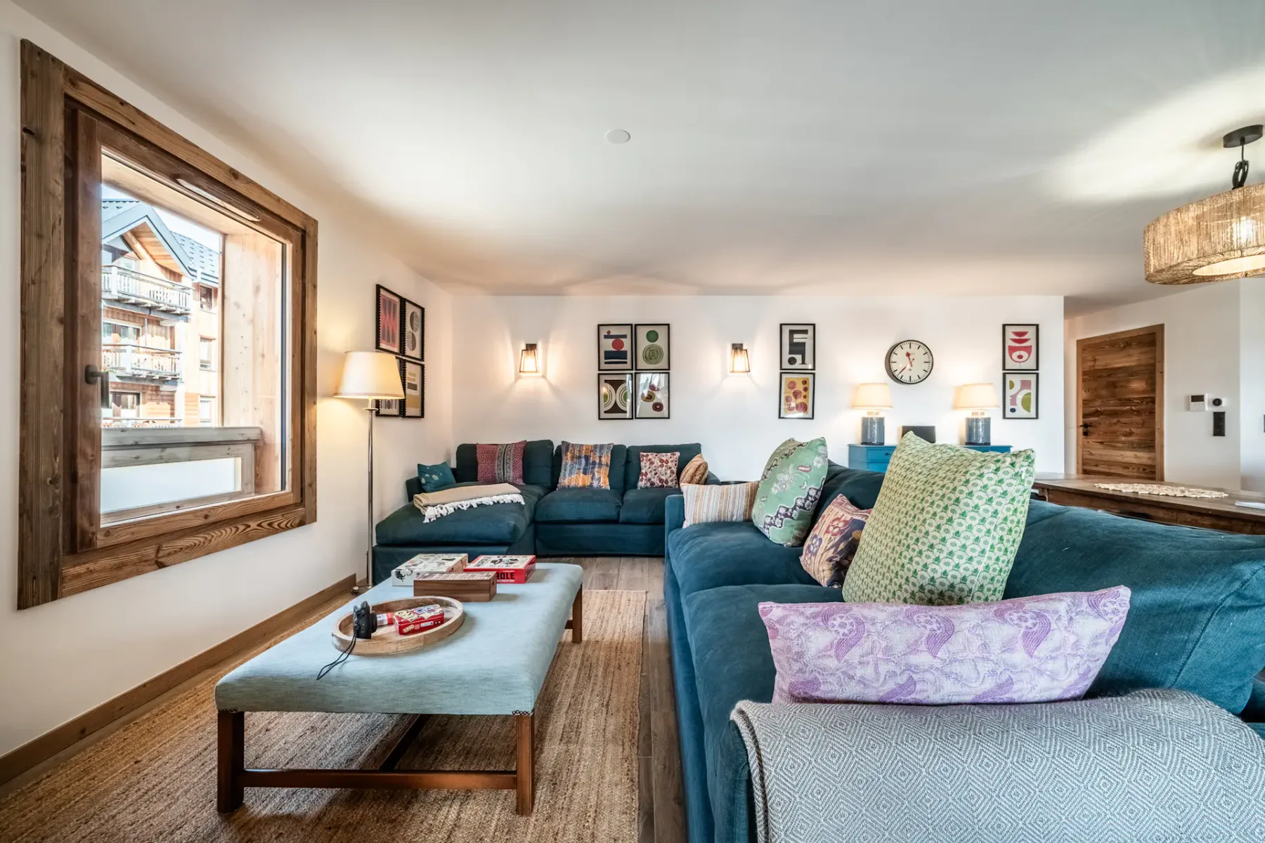 Living room with a teal sofa, orange armchair, and side tables. Wall art and lighting fixtures decorate white and wooden walls. A window provides natural light.