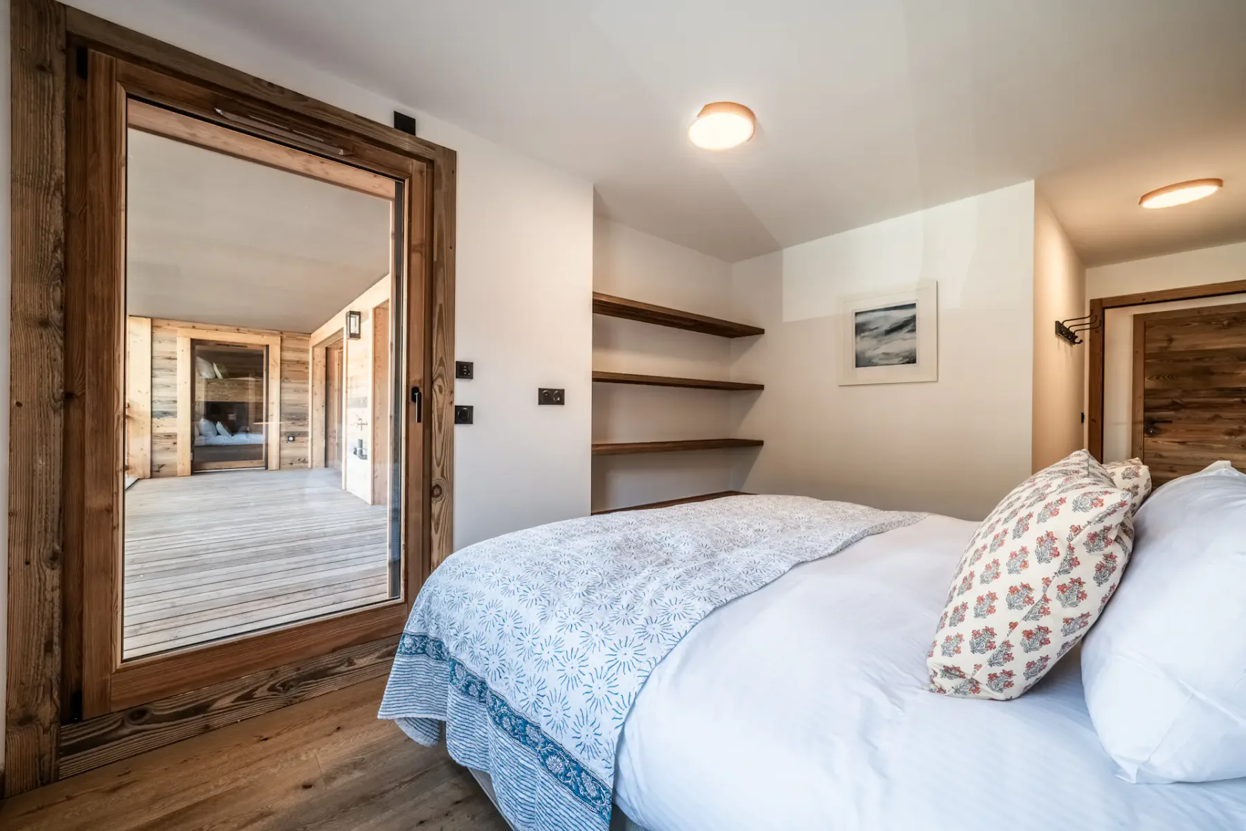 Cozy bedroom with a double bed, wooden walls, a bedside lamp, and built-in shelves. A window and glass door are visible, letting in natural light.