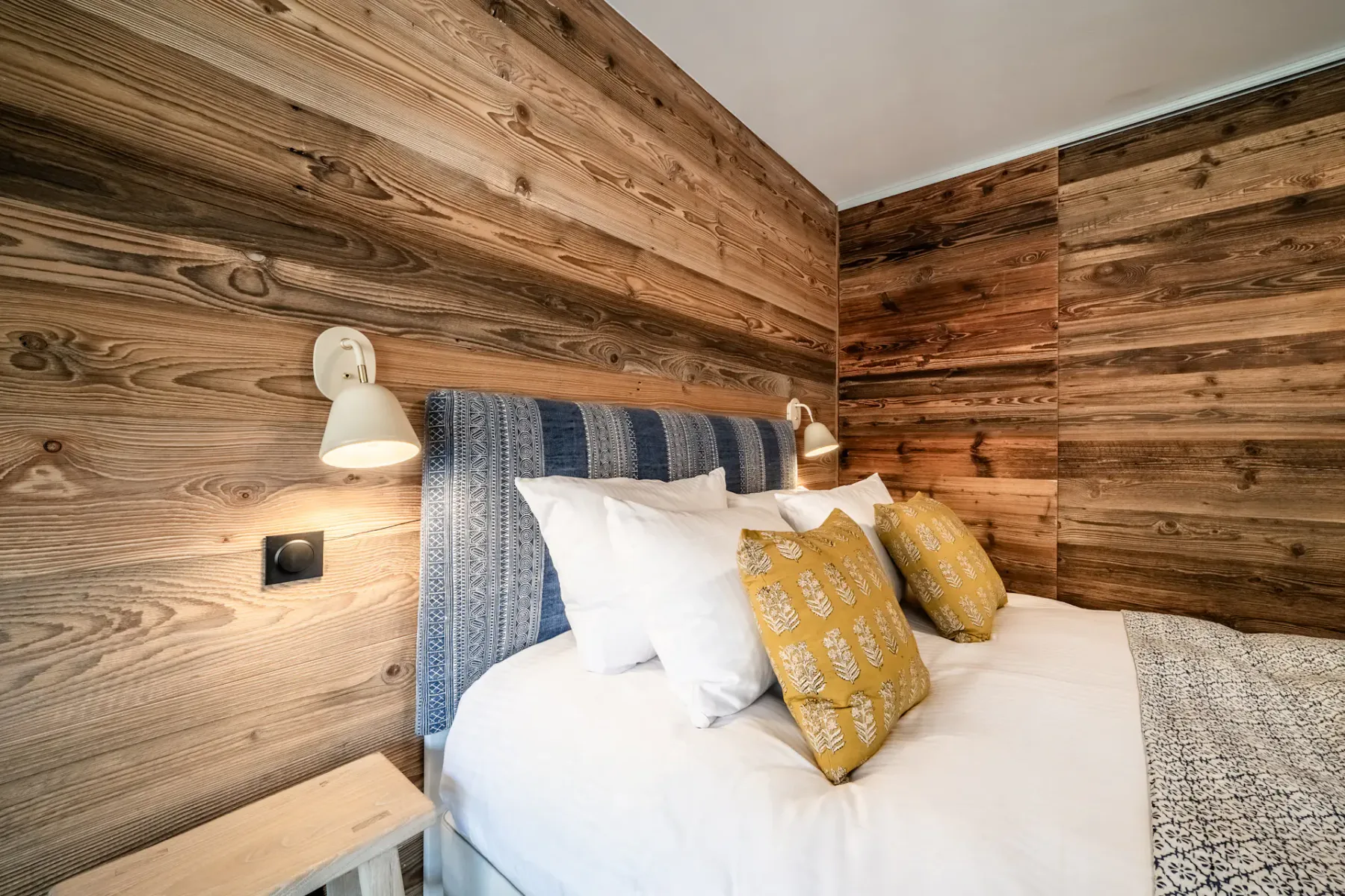 Cozy bedroom with a double bed, wooden walls, and large window doors opening to a balcony with a view of neighboring buildings.