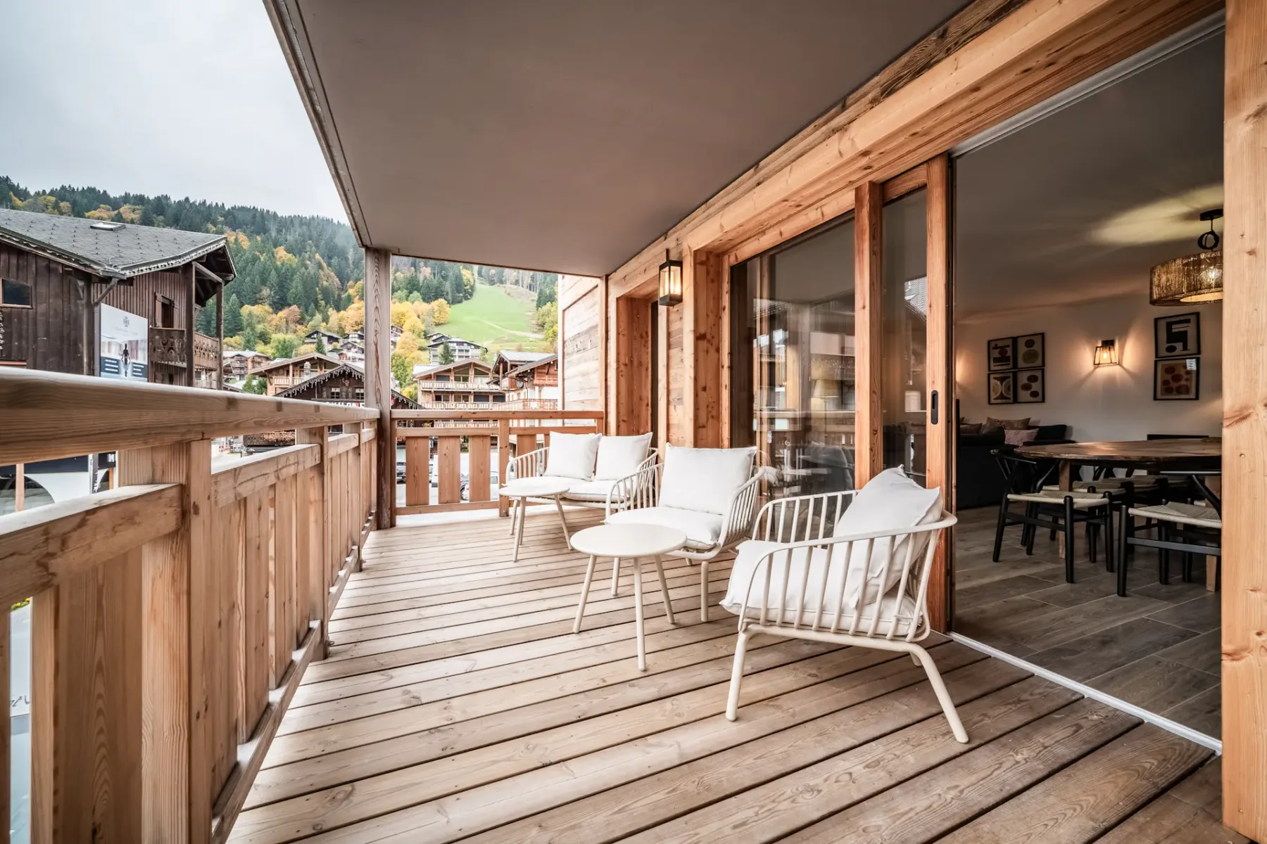 A wooden balcony with white chairs and small tables, overlooking neighboring buildings. Glass doors lead to the interior.