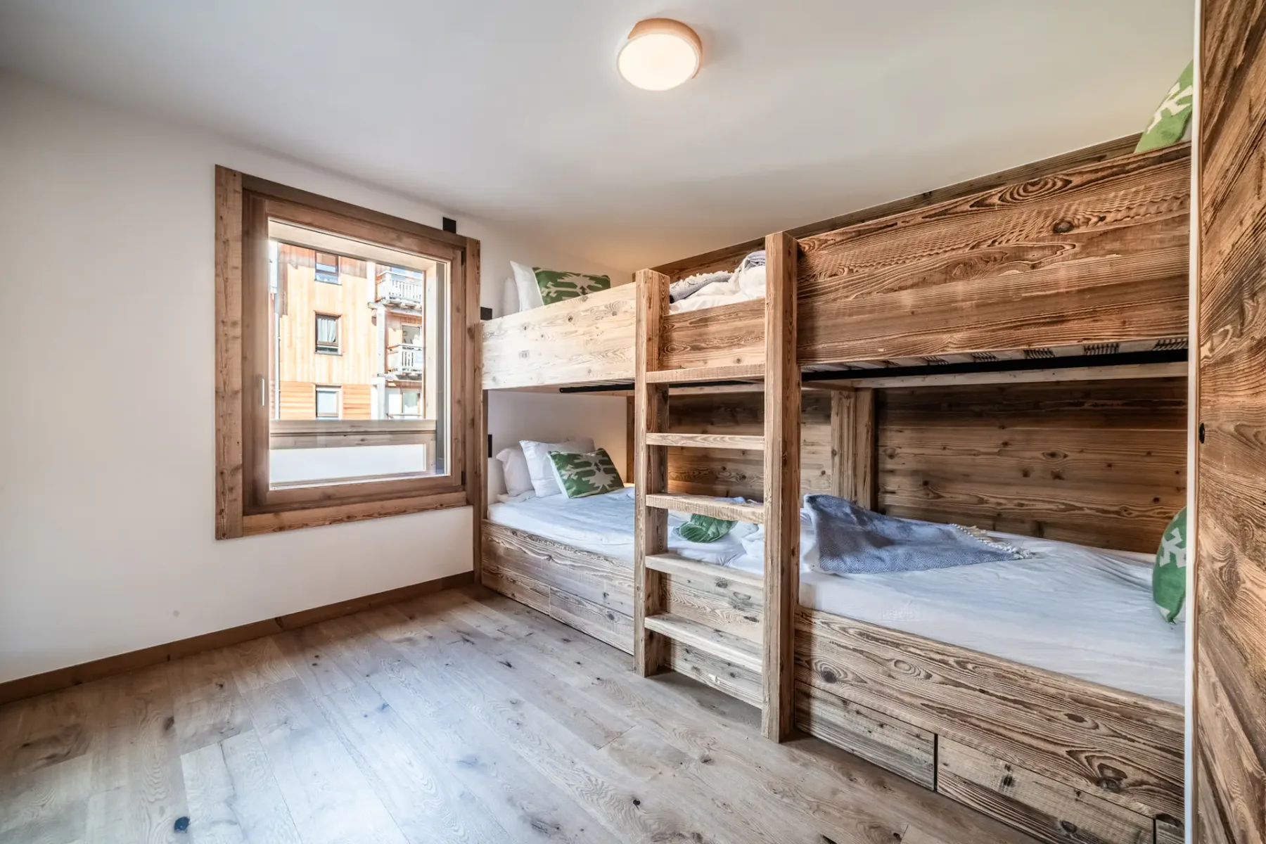 Wooden bunk beds in a bright room with a large window. The beds are neatly made with white linens. A ladder provides access to the upper bunks. The floor is wooden.