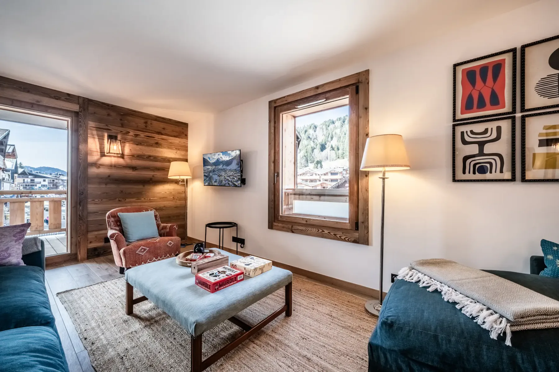 Living room with a teal sofa, orange armchair, and side tables. Wall art and lighting fixtures decorate white and wooden walls. A window provides natural light.