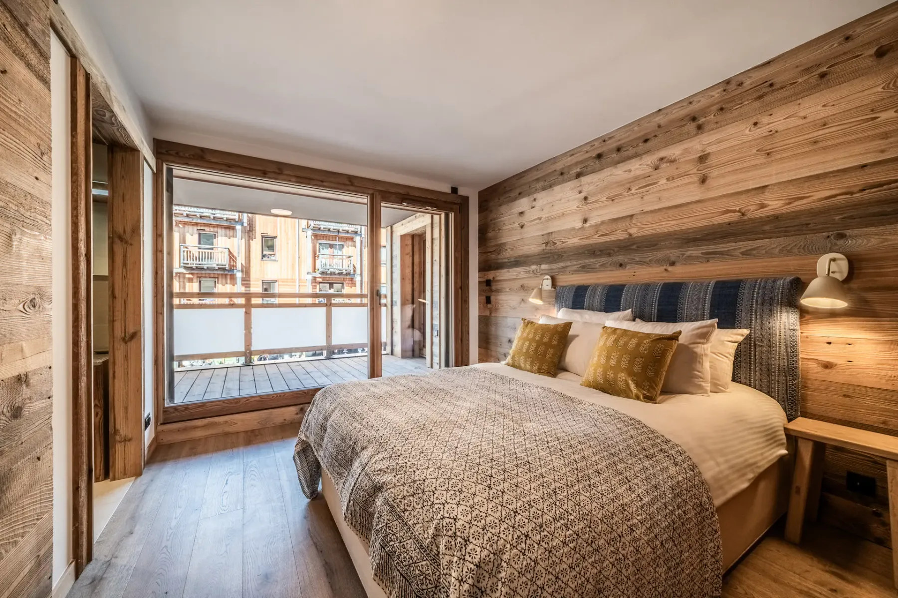 Cozy bedroom with a double bed, wooden walls, and large window doors opening to a balcony with a view of neighboring buildings.