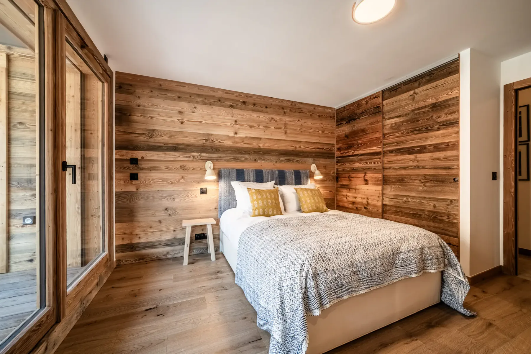 Cozy bedroom with a double bed, wooden walls, and large window doors opening to a balcony with a view of neighboring buildings.