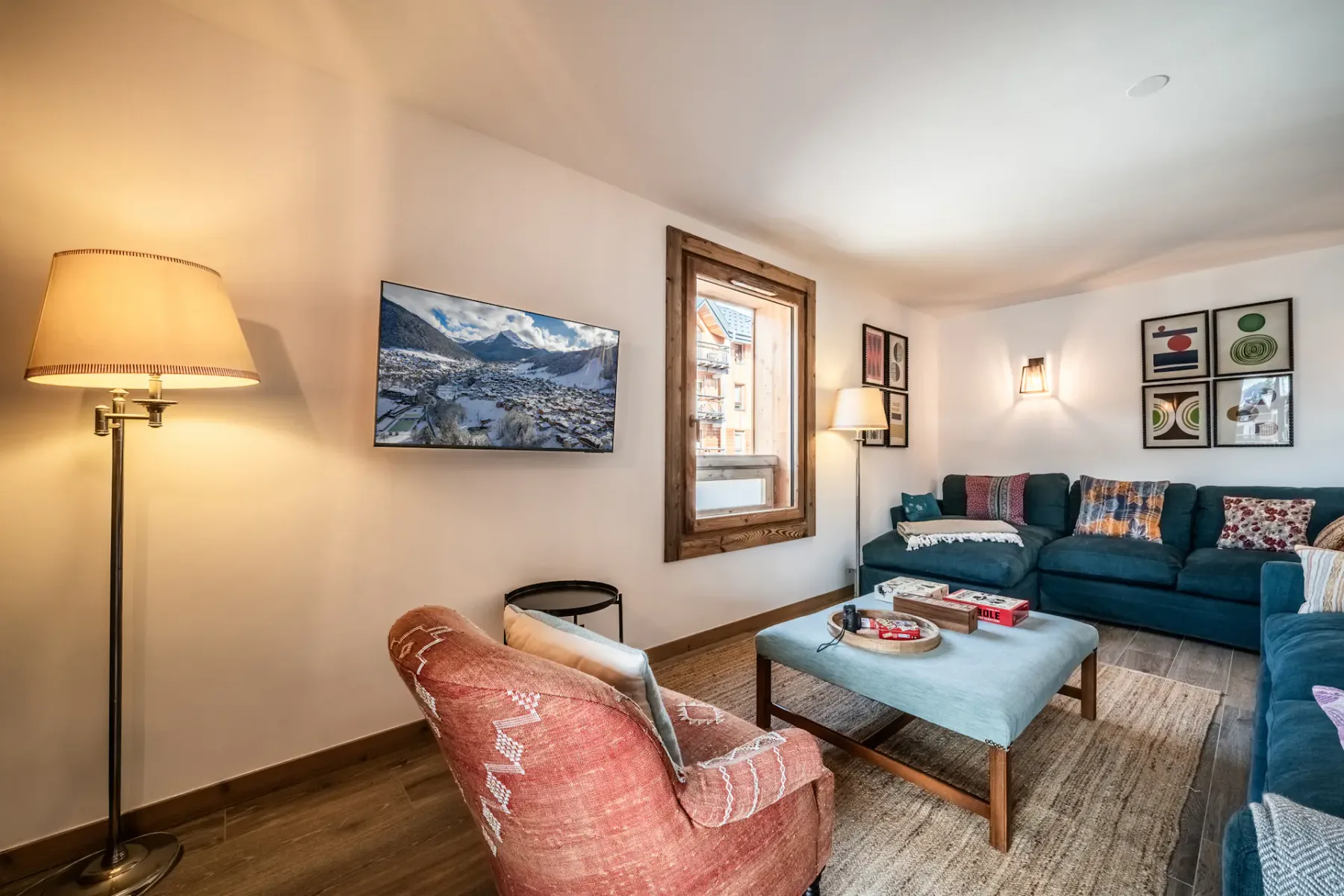 Living room with a teal sofa, orange armchair, and side tables. Wall art and lighting fixtures decorate white and wooden walls. A window provides natural light.