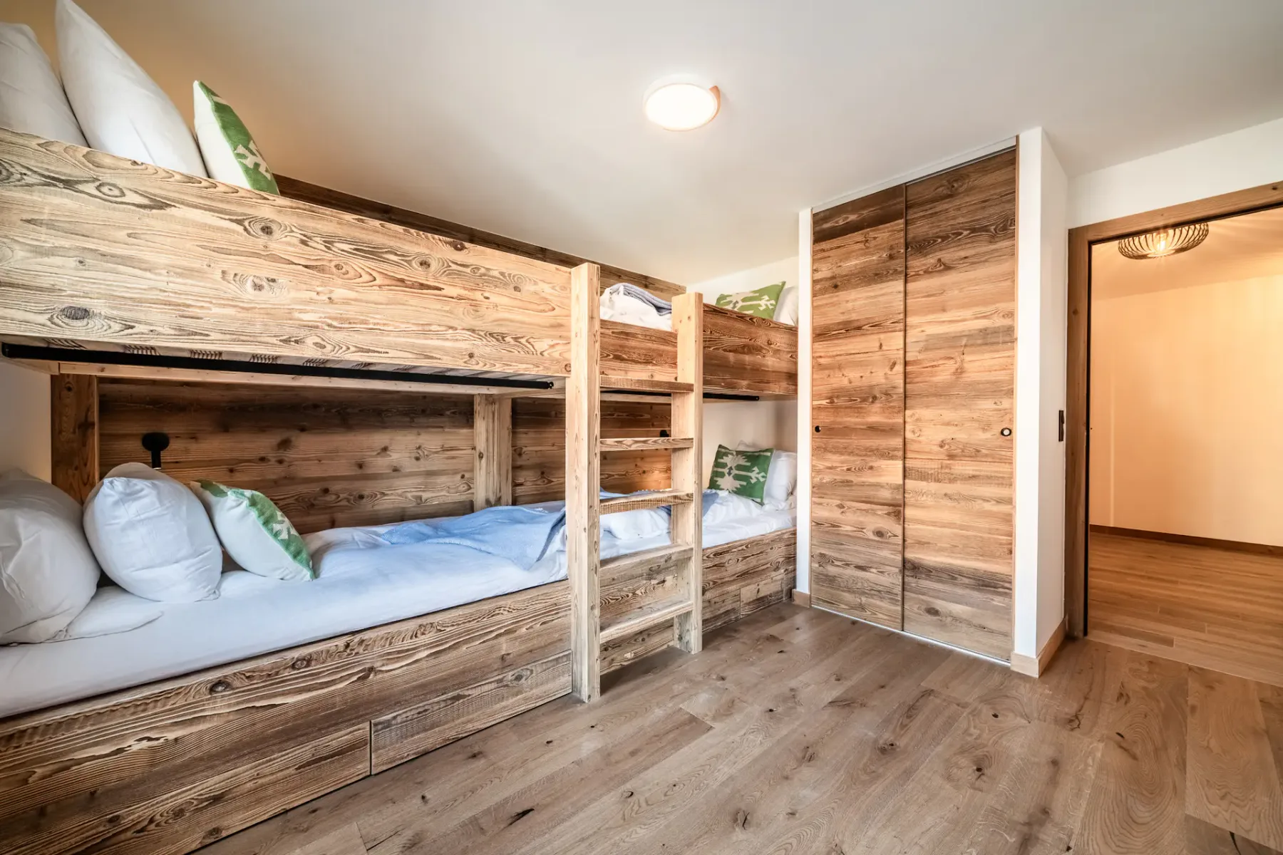 Wooden bunk beds in a bright room with a large window. The beds are neatly made with white linens. A ladder provides access to the upper bunks. The floor is wooden.