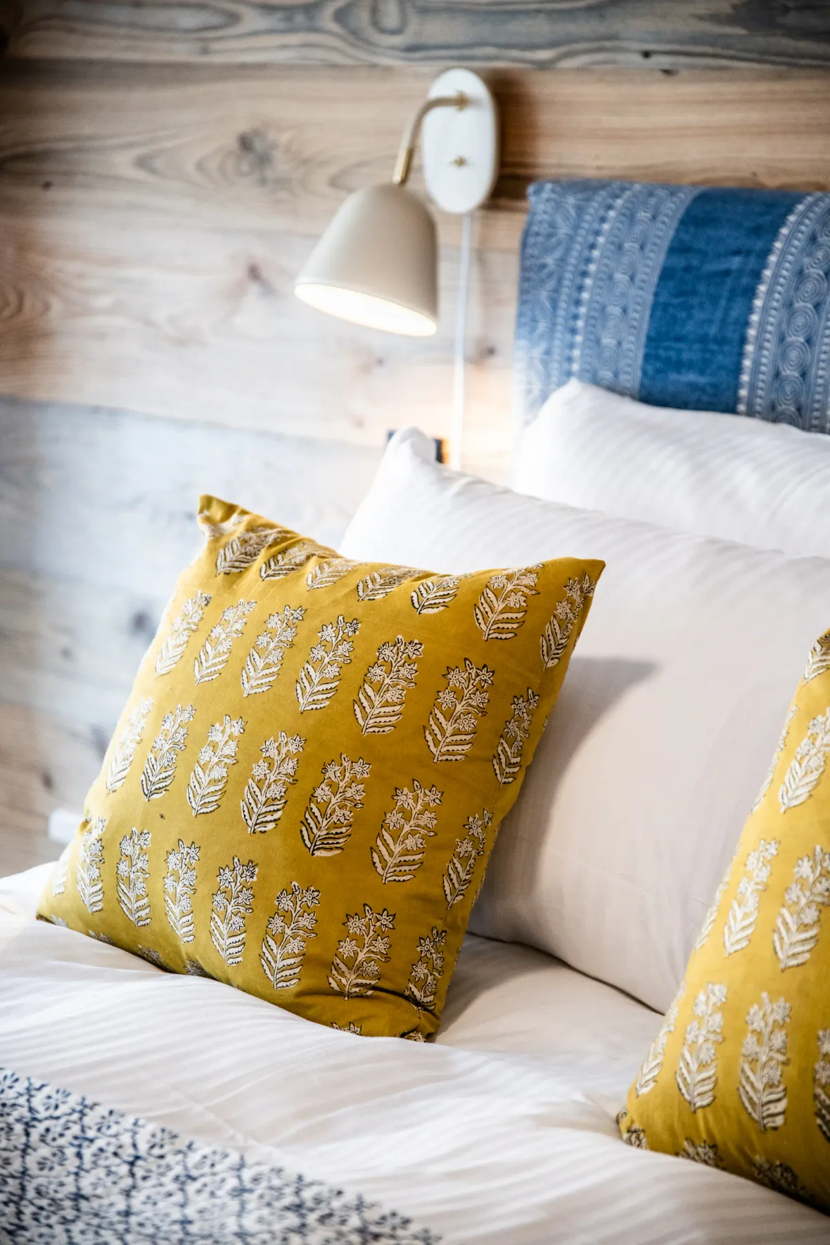 Close-up of a bed with white linens, two yellow decorative pillows with leaf patterns, and a wall-mounted reading lamp. A blue accent piece is seen at the headboard.