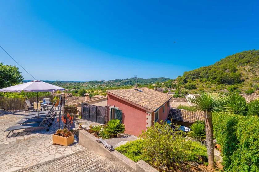 A stone terrace with outdoor seating overlooks a small house and green hills.