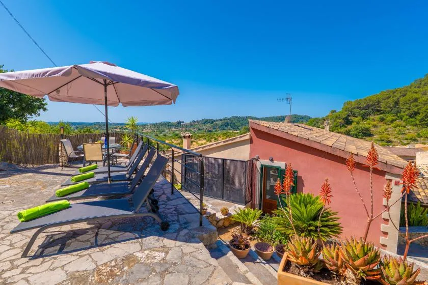 Una terraza soleada tiene asientos bajo una sombrilla y plantas junto al borde.