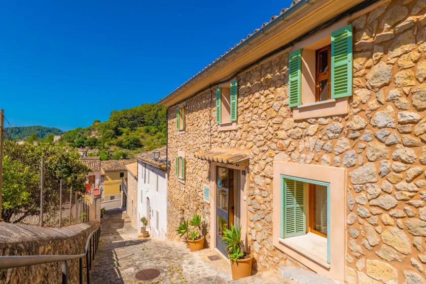 A stone house exterior with green shutters overlooks a narrow cobbled lane.