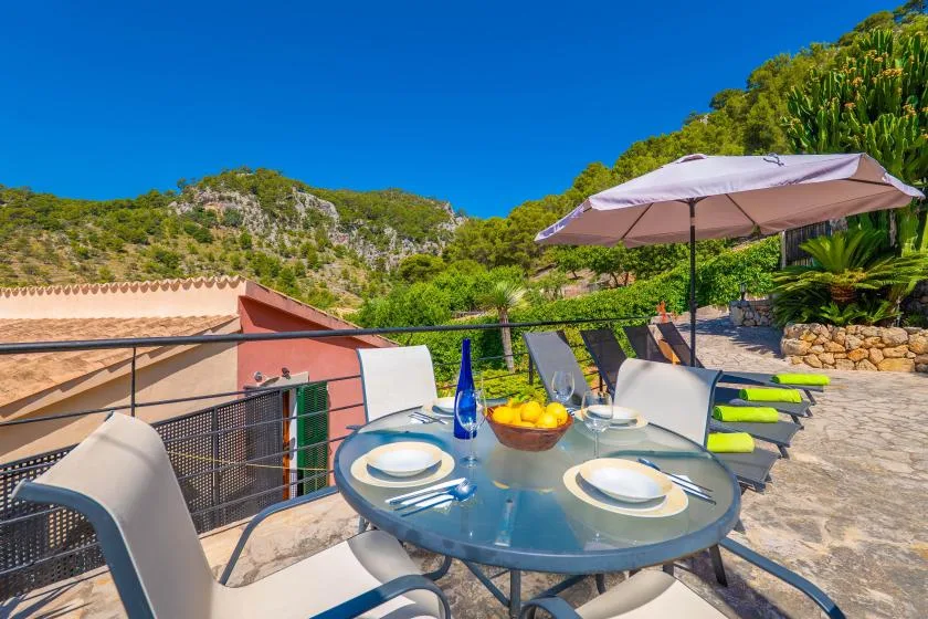 Una zona de comedor al aire libre con mesa redonda de vidrio y sombrilla mira hacia una colina verde.