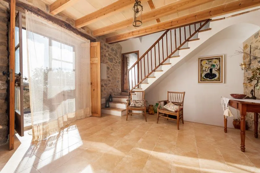 A bright living area has a wooden staircase, seating, and a large window.