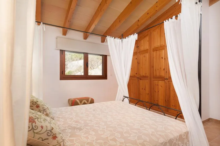 A bedroom with a bed, white curtains, and wooden ceiling beams.