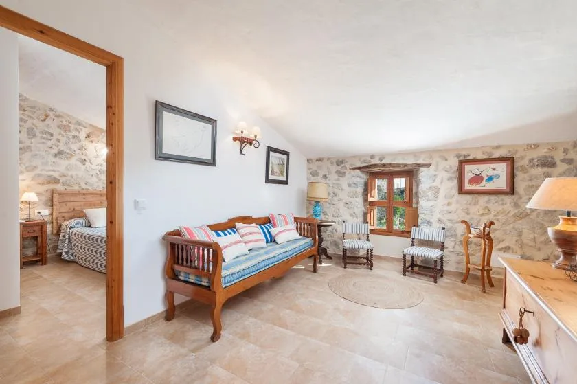 A bright living room with a wooden daybed, armchairs, and a stone wall.