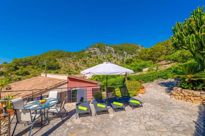 A sunny outdoor patio has seating beneath an umbrella, with green hills in the background.