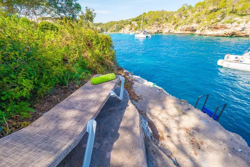A waterfront terrace overlooks bright blue water, with a lounge chair beside the shore.
