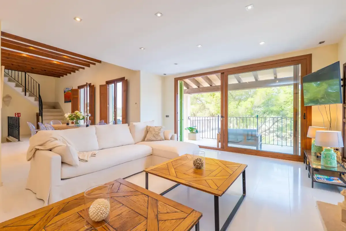 A bright living room with seating, wooden tables, and large windows opening onto a balcony.