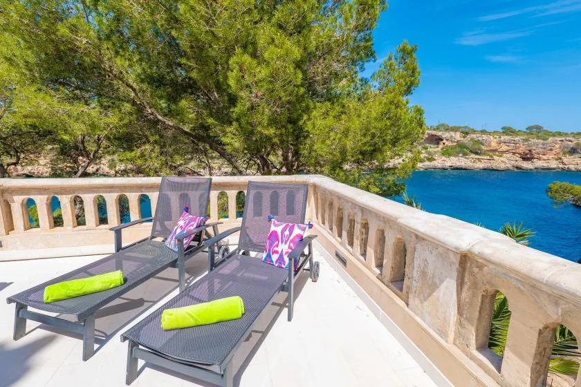 A sunny outdoor terrace overlooks the water, with lounge seating and a leafy tree.