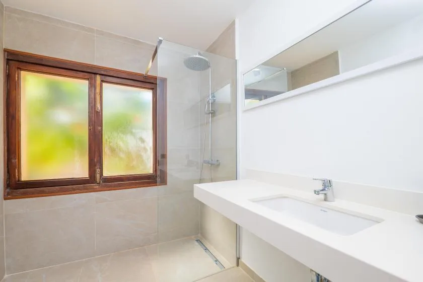 A bright bathroom has a long white sink counter and a glass shower beside a window.