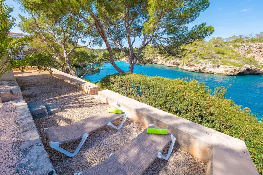 A seaside patio with reclining loungers overlooks bright blue water and a rocky coast.